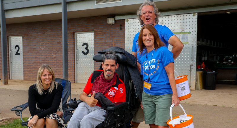 Miche, Sean, volunteers Deb and her husband Adrian are stood in front of the pavilion changing rooms. The volunteers are wearing the blue paul's place t-shirts with our logo and holding orange collection buckets. 