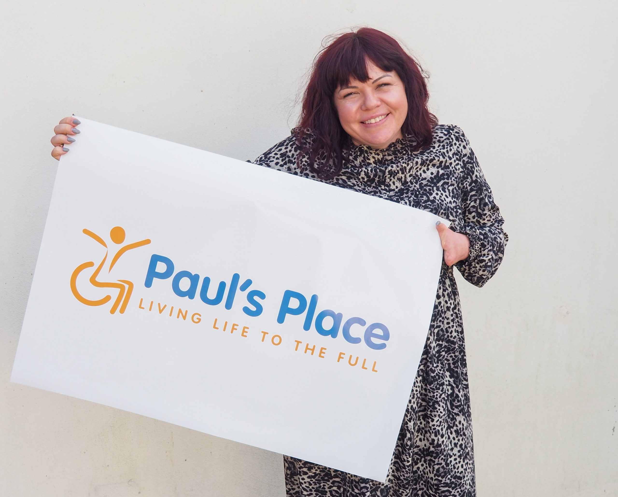 Briony May smiling holding up Paul's Place New Logo on a poster