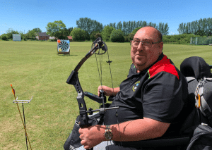 Andy Batten holding archery Bow and smiling. in the distance is a target